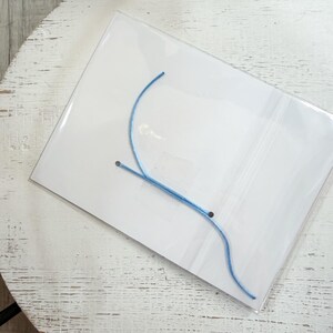 Op de afbeelding: Een doorzichtige plastic verpakking met een lichtblauw touwtje, mogelijk voor sieraden of knutselwerkjes. Het touwtje is gebogen en lijkt van een flexibel materiaal te zijn gemaakt. De verpakking ligt op een wit oppervlak met een verweerde afwerking.