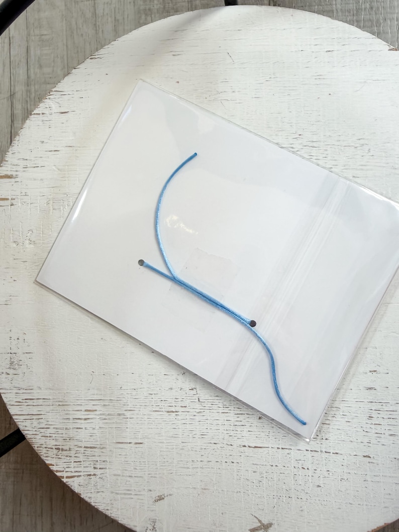 Op de afbeelding: Een doorzichtige plastic verpakking met een lichtblauw touwtje, mogelijk voor sieraden of knutselwerkjes. Het touwtje is gebogen en lijkt van een flexibel materiaal te zijn gemaakt. De verpakking ligt op een wit oppervlak met een verweerde afwerking.