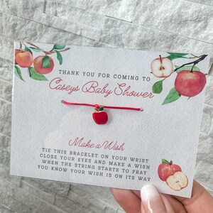 Puede incluir: Pulsera roja y verde con tem&aacute;tica de manzana con un cord&oacute;n rojo y un dije de manzana dorado. La tarjeta dice "Gracias por venir a la fiesta de bienvenida al beb&eacute; de Caseys. Pide un deseo. Ata esta pulsera en tu mu&ntilde;eca, cierra los ojos y pide un deseo. Cuando la cuerda empiece a deshilacharse, sabr&aacute;s que tu deseo est&aacute; en camino."
