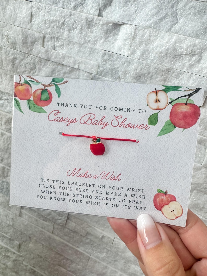 Puede incluir: Pulsera roja y verde con tem&aacute;tica de manzana con un cord&oacute;n rojo y un dije de manzana dorado. La tarjeta dice "Gracias por venir a la fiesta de bienvenida al beb&eacute; de Caseys. Pide un deseo. Ata esta pulsera en tu mu&ntilde;eca, cierra los ojos y pide un deseo. Cuando la cuerda empiece a deshilacharse, sabr&aacute;s que tu deseo est&aacute; en camino."