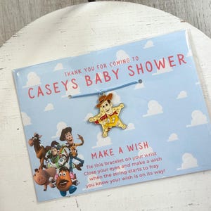 Op de afbeelding: Een babyshower gunst met een blauwe kaart met de tekst "CASEYS BABY SHOWER" en "MAKE A WISH". Het bevat een bedelarmband met een Woody-personageontwerp. De kaart heeft een lichtblauwe hemelachtergrond met witte wolken.