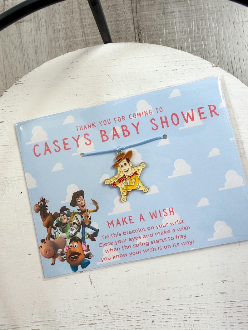Op de afbeelding: Een babyshower gunst met een blauwe kaart met de tekst "CASEYS BABY SHOWER" en "MAKE A WISH". Het bevat een bedelarmband met een Woody-personageontwerp. De kaart heeft een lichtblauwe hemelachtergrond met witte wolken.