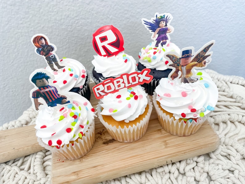 Puede incluir: Seis cupcakes decorados con chispas de colores y figuras de personajes de Roblox. Los cupcakes est&aacute;n sobre una tabla de cortar de madera. Las figuras incluyen un personaje de Roblox con un logotipo 'R' rojo, un personaje de Roblox con un logotipo 'Roblox' rojo, un personaje de Roblox con una camisa a rayas azul y roja, un personaje de Roblox con un vestido morado y alas, y un personaje de Roblox con una camisa amarilla y alas.