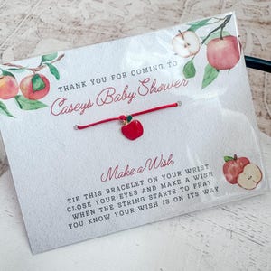 Puede incluir: Pulsera de cuerda roja con un peque&ntilde;o dije de manzana roja. La pulsera est&aacute; atada a una tarjeta blanca con el texto "Gracias por venir a la Baby Shower de Caseys" y "Pide un deseo". Ata esta pulsera en tu mu&ntilde;eca, cierra los ojos y pide un deseo. Cuando la cuerda empiece a deshilacharse, sabr&aacute;s que tu deseo est&aacute; en camino.