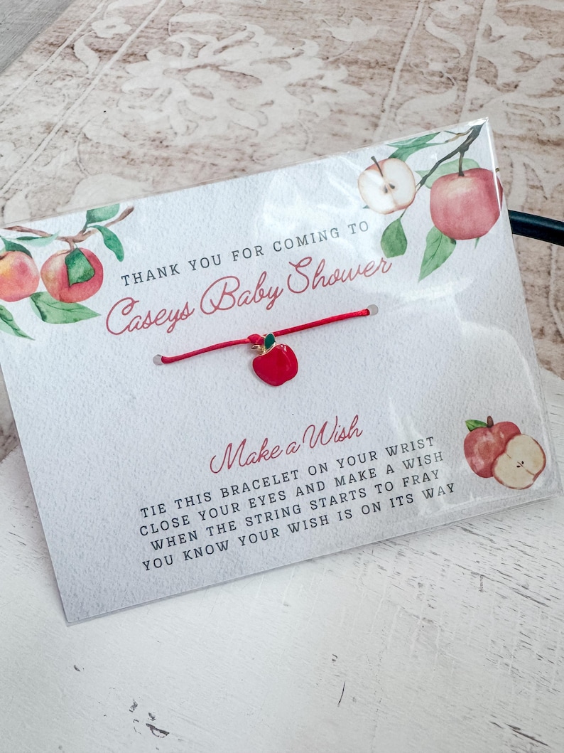 Puede incluir: Pulsera de cuerda roja con un peque&ntilde;o dije de manzana roja. La pulsera est&aacute; atada a una tarjeta blanca con el texto "Gracias por venir a la Baby Shower de Caseys" y "Pide un deseo". Ata esta pulsera en tu mu&ntilde;eca, cierra los ojos y pide un deseo. Cuando la cuerda empiece a deshilacharse, sabr&aacute;s que tu deseo est&aacute; en camino.