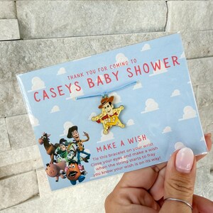 Op de afbeelding: Een baby shower gunst met een blauwe armband met een Woody bedel. De kaart leest "Bedankt voor je komst naar Caseys Baby Shower" met een "Doe een wens" bericht en Toy Story karakters.