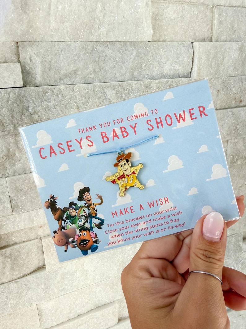 Op de afbeelding: Een baby shower gunst met een blauwe armband met een Woody bedel. De kaart leest "Bedankt voor je komst naar Caseys Baby Shower" met een "Doe een wens" bericht en Toy Story karakters.