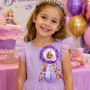 Op de afbeelding: Een lichtpaarse jurk met ruches van tule mouwen en een ronde halslijn. Een ronde paarse rozetbadge is op de borst gespeld, met de tekst Birthday Girl en een illustratie van een personage met lang blond haar in een paarse jurk.