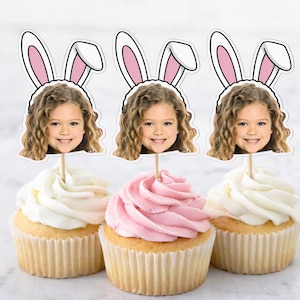 Puede incluir: Tres cupcakes con glaseado blanco y rosa, cada uno coronado con una foto de un niño sonriente con orejas de conejo. Los cupcakes están sobre una superficie blanca, perfectos para una celebración de Pascua o una fiesta infantil.
