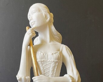 May include: A white porcelain figurine of a woman holding a gold-painted broom. She is wearing a white dress with a gold-painted bodice. The figurine is standing on a white base with a decorative design.