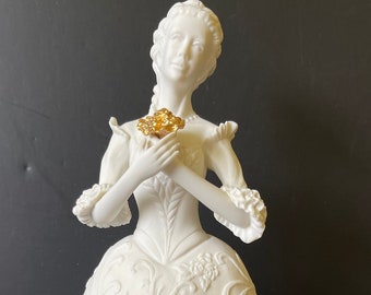 May include: A white porcelain figurine of a woman in a flowing gown, holding a gold flower. The gown is decorated with floral patterns and the figurine is standing on a white base.