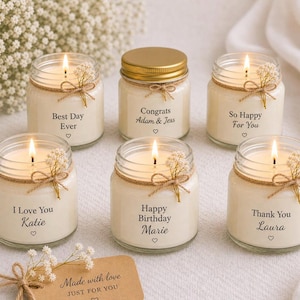 May include: Six lit, small glass jar candles with various messages, including "Best Day Ever," "Congrats Adam & Jess," and "Happy Birthday Marie." Each candle is decorated with twine, dried flowers, and a heart symbol. A small card reads "Made with love."