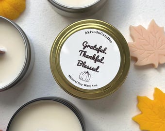 May include: A collection of soy wax candles in silver tins, one with a gold lid featuring the text "Grateful. Thankful. Blessed." and a pumpkin graphic. The candles are surrounded by fall-themed decorations, including orange and peach-colored leaf and pumpkin shapes.