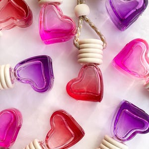 May include: Eight heart-shaped glass bottles with a gradient of pink, purple, and red colors. Each bottle has a wooden ring and a string for hanging. The bottles are on a white surface.