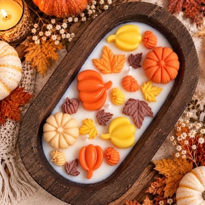 May include: A dark brown wooden dough bowl candle filled with white wax and topped with various wax embeds shaped like orange, yellow, and cream pumpkins and autumn leaves. The candle features multiple wicks embedded within the wax surface.