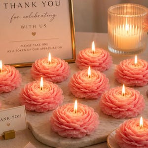 K&ouml;nnte beinhalten: Mehrere rosa blumenf&ouml;rmige Kerzen brennen auf einer Marmoroberfl&auml;che. Jede Kerze hat geschichtete Bl&uuml;tenbl&auml;tter und eine einzelne Flamme. Ein Schild im Hintergrund zeigt den Text, THANK YOU for celebrating WITH US, PLEASE TAKE ONE AS A TOKEN OF OUR APPRECIATION.
