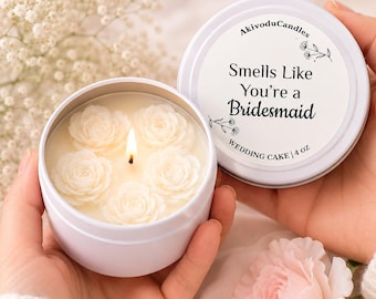 Puede incluir: Una vela en lata blanca con una mecha encendida rodeada de cinco pequeñas rosas de cera blanca. La tapa presenta el texto AkivoduCandles, Smells Like You're a Bridesmaid, WEDDING CAKE y 4 oz. La vela se sostiene en una mano.