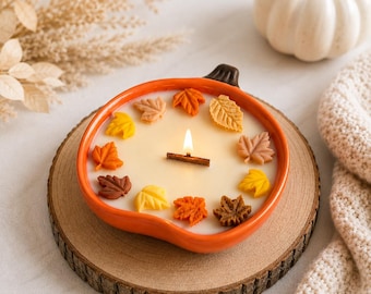 May include: A pumpkin-shaped ceramic candle holder with a glossy orange finish, containing white wax and a wooden wick. The candle is decorated with small, colourful wax leaves in shades of yellow, orange, and brown arranged on the surface.