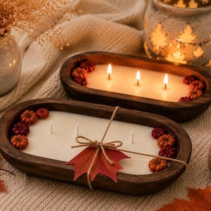 May include: Two oval-shaped wooden dough bowls filled with white wax and three wicks each. The candles are decorated with small orange and burgundy pumpkins. One bowl is tied with twine and features a single red maple leaf paper tag.