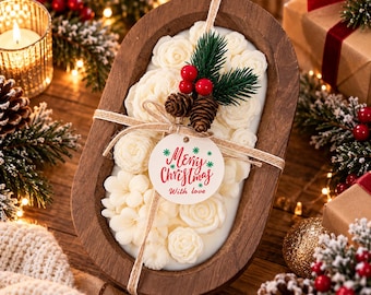 Könnte beinhalten: Eine ovale Holzschale gefüllt mit weißen Wachsblumen, verziert mit einem Zweig aus grünen Tannennadeln, roten Beeren und zwei kleinen Kiefernzapfen. Eine beigefarbene Kordel hält ein rundes weißes Etikett mit der Aufschrift Merry Christmas With love.