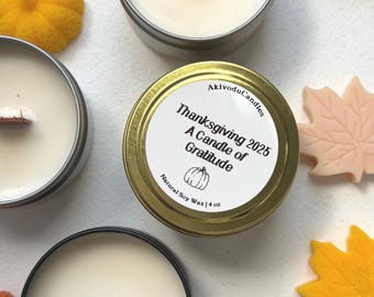 May include: A close-up of several small, round candles in metal tins. The central candle has a label that reads "Thanksgiving 2025 A Candle of Gratitude" with a pumpkin graphic. The candles are surrounded by fall-themed decorations.
