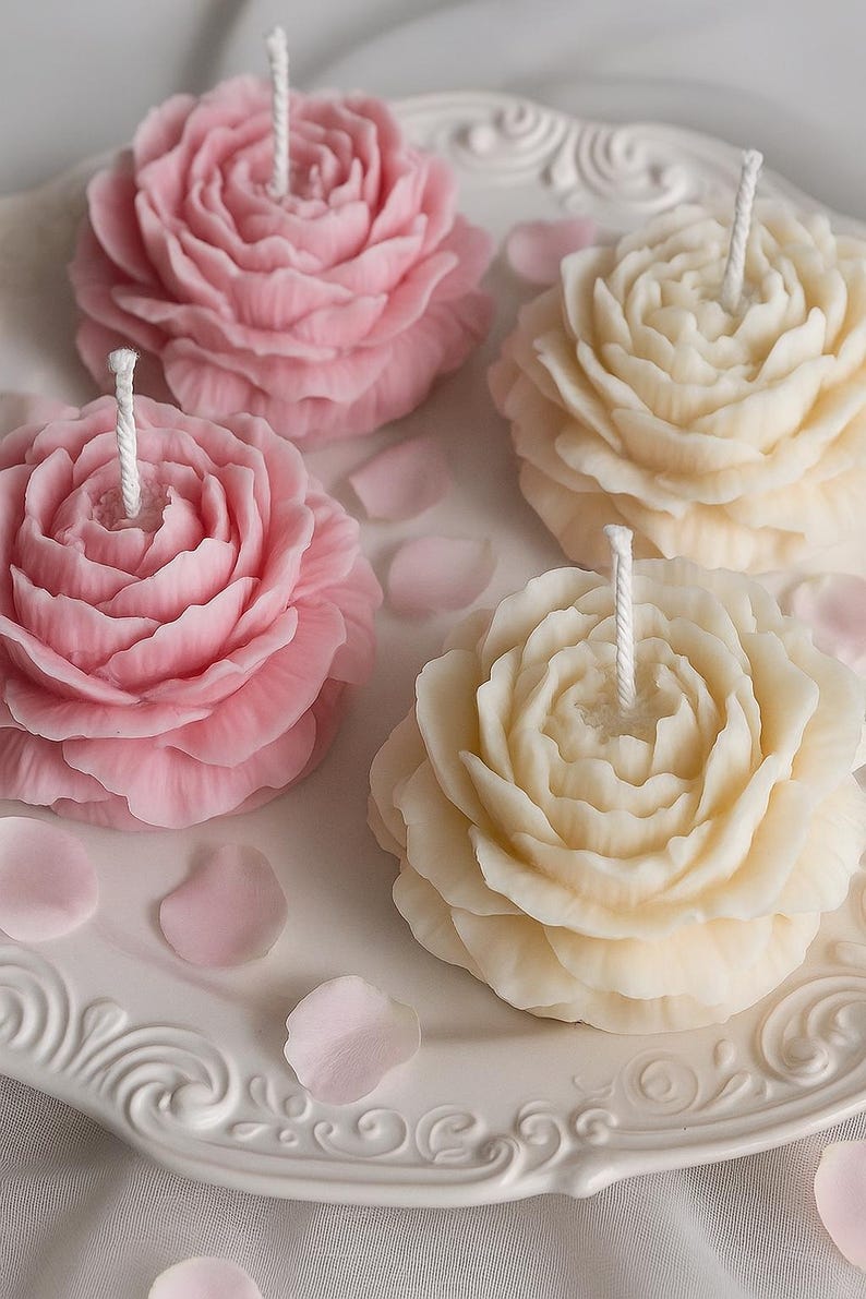 K&ouml;nnte beinhalten: Vier blumenf&ouml;rmige Kerzen auf einem wei&szlig;en Zierteller. Zwei Kerzen sind hellrosa und zwei cremefarben, jede mit geschichteten Bl&uuml;tenbl&auml;ttern und einem zentralen wei&szlig;en Docht. Kleine, lose rosa Bl&uuml;tenblattfragmente sind auf dem Teller verstreut.