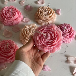 K&ouml;nnte beinhalten: Eine Sammlung blumenf&ouml;rmiger Kerzen in Rosa- und Beiget&ouml;nen, jede mit detaillierten Bl&uuml;tenbl&auml;ttern und einem zentralen Docht. Die Kerzen sind zusammen mit verstreuten Bl&uuml;tenbl&auml;ttern angeordnet, wobei eine rosa Kerze im Vordergrund gehalten wird.