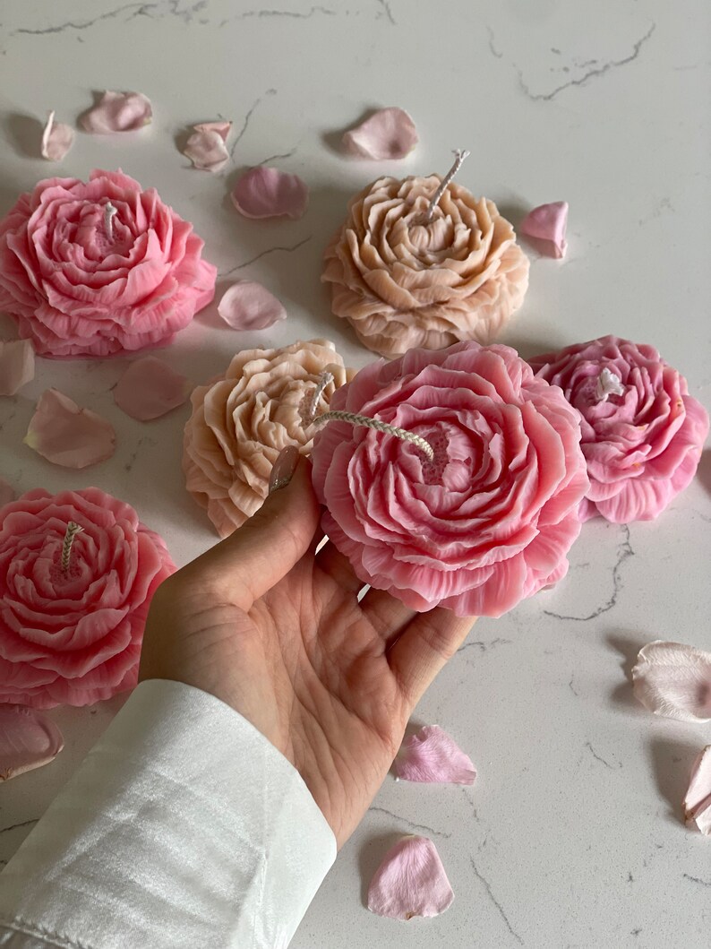 K&ouml;nnte beinhalten: Eine Sammlung blumenf&ouml;rmiger Kerzen in Rosa- und Beiget&ouml;nen, jede mit detaillierten Bl&uuml;tenbl&auml;ttern und einem zentralen Docht. Die Kerzen sind zusammen mit verstreuten Bl&uuml;tenbl&auml;ttern angeordnet, wobei eine rosa Kerze im Vordergrund gehalten wird.