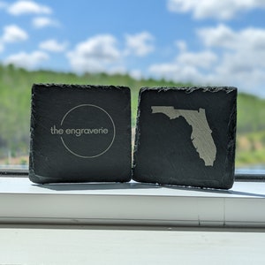 May include: Two black slate coasters. One coaster features the text "the engravmerie" in a circle. The other coaster features a white outline of the state of Florida.