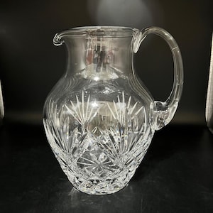 May include: A clear glass pitcher with a cut crystal design. The pitcher has a wide base and a narrow neck.