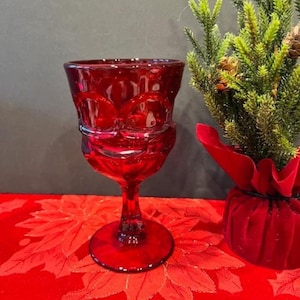 Puede incluir: Una copa de vidrio de color rojo intenso con base con tallo. La copa tiene un diseño texturizado y está sobre un mantel rojo con estampado floral.