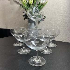 May include: Set of six clear glass stemmed wine glasses with a beaded design on the stem. The glasses are arranged in a circle on a black surface.