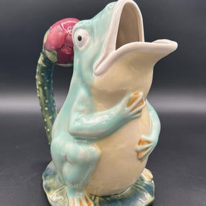 May include: A ceramic frog-shaped pitcher. The pitcher is light blue with a cream belly and orange feet. The handle is a green cactus with a red flower. The frog sits on a green lily pad. Measurements are in centimeters.