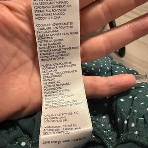 May include: A white tag with black text detailing the fabric composition and care instructions for a green and white polka dot dress. The tag includes the brand name "Urban Outfitters, Inc." and the address "146 Brick Lane, London, E1 6RU".