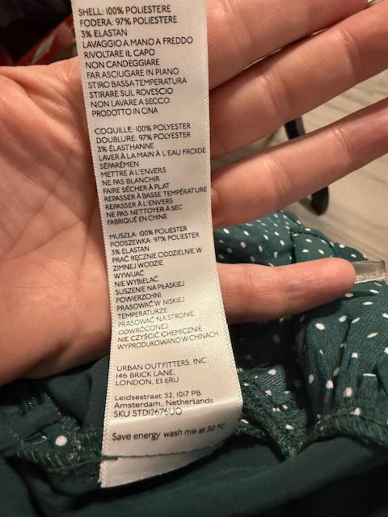 May include: A white tag with black text detailing the fabric composition and care instructions for a green and white polka dot dress. The tag includes the brand name "Urban Outfitters, Inc." and the address "146 Brick Lane, London, E1 6RU".