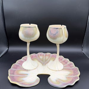 May include: A set of two ceramic tulip-shaped wine glasses with a matching leaf-shaped tray. The glasses and tray are off-white with pink, gold, and purple accents. The tray has a scalloped edge and space for the glasses.