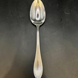 Puede incluir: Una cuchara plateada con un cuenco ovalado y un mango detallado. El mango presenta un diseño acanalado vertical con un borde decorativo. La cuchara está hecha de un metal brillante y reflectante, probablemente acero inoxidable o plata.