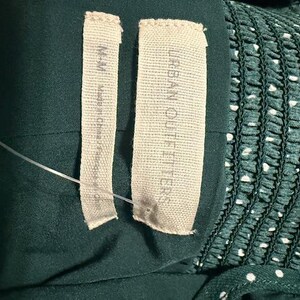 May include: Close-up of two white fabric labels sewn onto a dark green garment. The label on the left says "Made in China". The label on the right says "Urban Outfitters".
