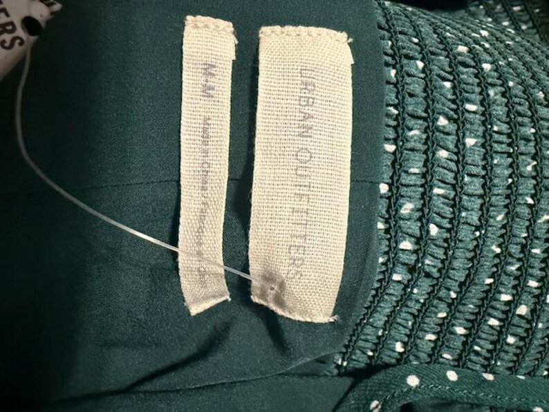 May include: Close-up of two white fabric labels sewn onto a dark green garment. The label on the left says "Made in China". The label on the right says "Urban Outfitters".