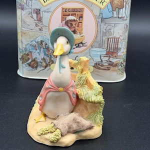 May include: A ceramic figurine of a goose wearing a blue bonnet and pink shawl, leaning against a wooden fence. A sack of grain sits at its feet. The figurine is in front of a tin with "The World of Beatrix Potter" printed on it.