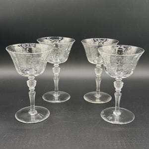 Puede incluir: Cuatro copas de cóctel transparentes con diseños florales grabados. Cada copa tiene una base redonda, un tallo detallado y un cuenco ancho y poco profundo. Las copas están dispuestas sobre una superficie oscura.