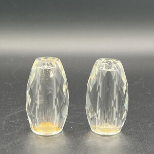 Può includere: Due oggetti di vetro trasparente sfaccettati con un motivo a diamante. Gli oggetti sono di forma ovale con una piccola apertura nella parte superiore e una base leggermente giallastra. Sono posti su uno sfondo grigio scuro.