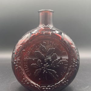 May include: A ruby red glass bottle with a round body and a short neck. The bottle features embossed text and a holly leaf design. The text reads "CHRISTMAS HOLLY 1876".
