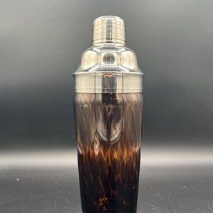Pode incluir: Uma coqueteleira com tampa e base de aço inoxidável. O corpo de vidro tem um padrão manchado marrom e âmbar. A coqueteleira foi projetada para misturar bebidas e tem uma aparência clássica e elegante.