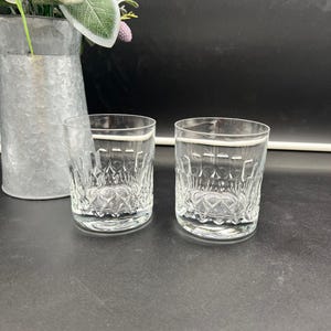May include: Two clear glass tumblers with a cut-glass design. The glasses are empty and sitting on a dark surface.