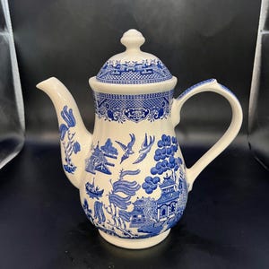 Puede incluir: Una tetera de cerámica blanca y azul con tapa. La tetera presenta un diseño clásico de patrón Willow con ilustraciones azules de pagodas, árboles y pájaros. El asa y el pico son blancos.