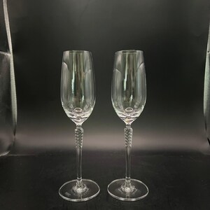 May include: A pair of clear champagne flutes with a twisted stem design. The flutes have a long, slender stem and a wide, rounded bowl. The base is a flat, circular design. The glasses are made of clear glass against a black background.