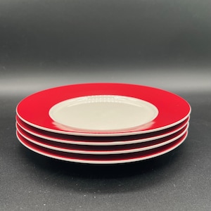 May include: A stack of four ceramic plates. Each plate has a wide red rim and a white center. The plates are stacked on top of each other, with the top plate showing the full design. The plates are on a dark background.