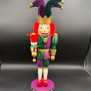 May include: A decorative wooden nutcracker in the form of a jester. The jester wears a purple and green hat, a gold crown, and a red wig. The jester's outfit is green and purple with gold accents. The nutcracker stands on a purple base with a gold and green diamond pattern.