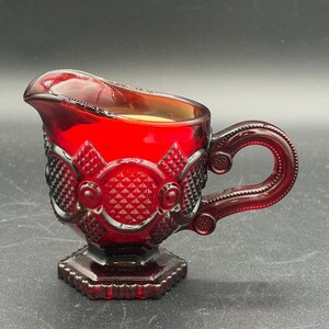 May include: A deep red glass creamer pitcher featuring a textured diamond pattern and circular motifs on the exterior. The vessel has a curved handle, a wide pouring spout, and a hexagonal base, showcasing a translucent finish throughout the glass.